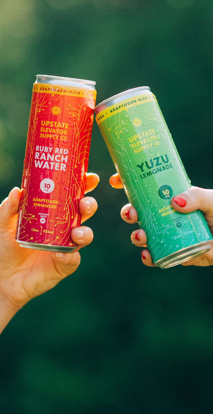 Two hands holding a Ruby Red Ranch Water and Yuzu Lemonade cans against a blurred natural background
